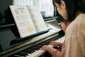 「ピアノの前に座り、母に見守られながら自力で楽譜を読み取ろうとする子供。初見演奏による自立と脳トレをイメージした写真」