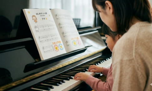 「ピアノの前に座り、母に見守られながら自力で楽譜を読み取ろうとする子供。初見演奏による自立と脳トレをイメージした写真」
